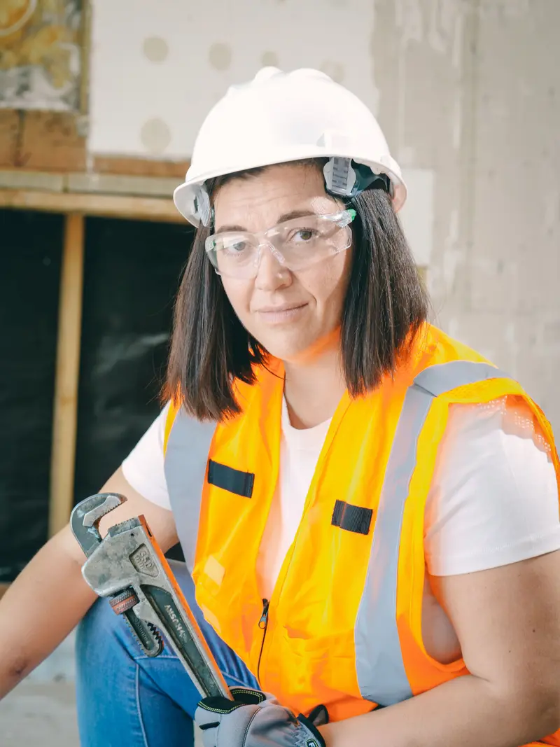 A smiling tradeswoman in a hard hat and orange hi-vis holding a wrench